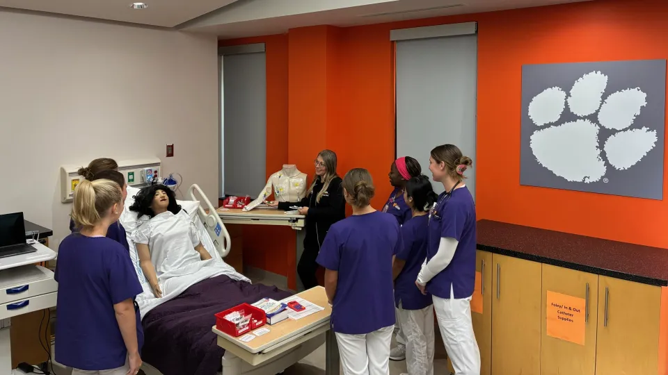 One professor in black and six students in purple and white scrubs in a nursing clinical lab.